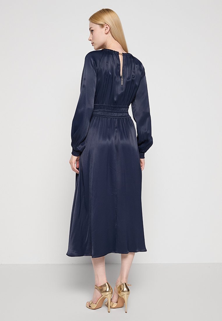 Abito di raso blu navy con maniche lunghe, vita elasticizzata, apertura a chiave sul retro e orlo di lunghezza media. Completato da tacchi a strappo dorati.
