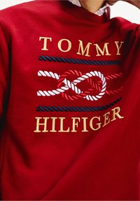 Red sweatshirt featuring "TOMMY HILFIGER" in gold embroidery, with a nautical rope design in red, white, and blue across the chest.