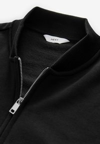 Veste zippée noire avec un col côtelé et une texture douce. Présente une fermeture éclair métallique et une étiquette avec le label "NEXT" à l'intérieur du col.