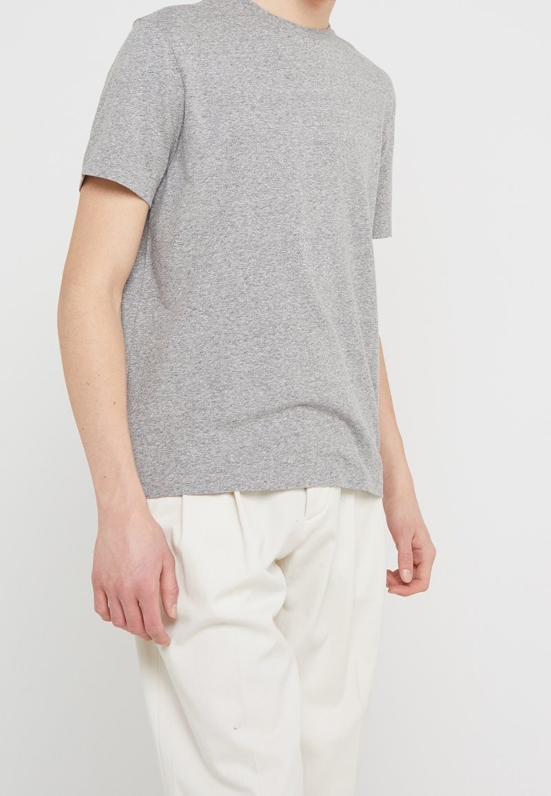 Homme portant un t-shirt gris uni à manches courtes et un pantalon blanc ample, debout devant un fond neutre.