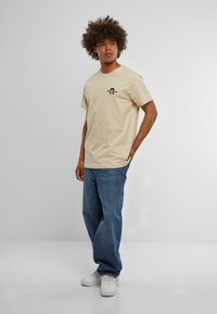 Beige cotton crew neck t-shirt featuring a black graphic print, paired with loose denim jeans and white sneakers.