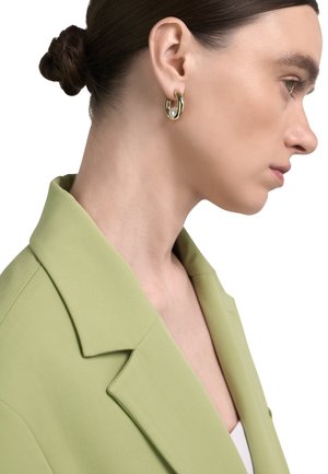 Tumejuustega naine, kes kannab kuldseid kõrvarõngaid pärliga ja helerohelist bleiserit, vaadates paremale valgel taustal.