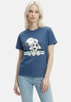 Junge Frau mit langen blonden Haaren, die ein blaues T-Shirt mit Snoopy, der einen Fußball hält, trägt, kombiniert mit hellblauen Jeans.