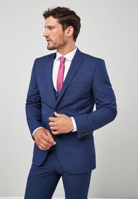 Marineblaues Sakko mit schlanker Passform, einreihigem Design und Zwei-Knopf-Verschluss. Ausgestattet mit einem weißen Hemd und einer rosa Krawatte sowie Seitentaschen.