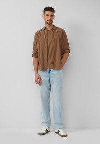 Braun gestreiftes Hemd mit Knopfleiste, kombiniert mit hellblauen gerade geschnittenen Jeans und weißen Sneakers mit dunklen Akzenten. Einfarbiger Hintergrund.