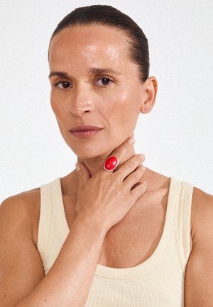 Naine kreemika tankitopiga, juuksed tagasi kammitud, käega kaela hellitamas, kannab keskmisel sõrmel suurt punast ovaalset sõrmust.