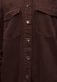 Gros plan d'une chemise marron foncé avec deux poches à rabat sur la poitrine et cinq boutons-pression métalliques sur le devant.