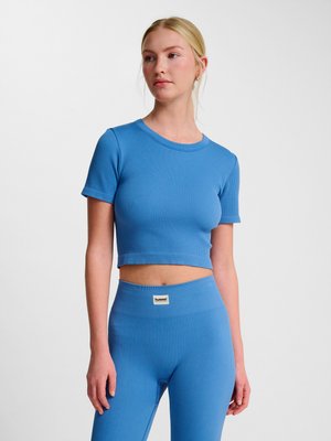 Ung kvinde iført matchende blå ribstrikket crop top og højtaljede leggings står mod en ens hvid baggrund.