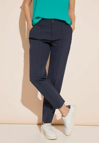 Pantaloni tailleur blu navy con gamba dritta, dotati di tasche frontali e chiusura con bottone. Abbinati a sneakers bianche e una maglia verde.