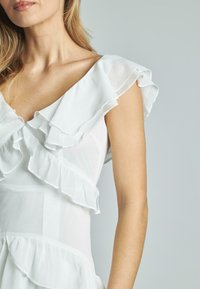 Weißes Kleid mit gestuften Rüschen am Ausschnitt und den Ärmeln, aus einem strukturierten Stoff. Das Design umfasst ein tailliertes Oberteil und einen weit auslaufenden Saum.