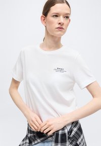 Νέα γυναίκα με ανοιχτόχρωμο δέρμα και καστανά μαλλιά φοράει ένα λευκό T-shirt της GANT και δένει ένα ασπρόμαυρο καρό πουκάμισο γύρω από τη μέση της.