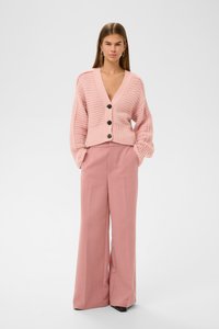 Růžový pletený cardigan s hlubokým výstřihem do V, oversized rukávy a černými knoflíky, doplněný o růžové kalhoty vysokého pasu s širokými nohavicemi.