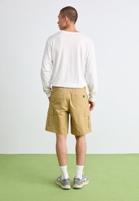 Bermudas cargo beiges com múltiplos bolsos, combinadas com uma camisa de manga longa branca. O visual inclui ténis cinzentos e meias brancas até ao tornozelo.