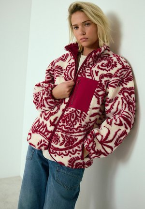 Fleece jacket with cream and red floral pattern, high collar, front zipper, and a single maroon pocket. The model wears denim pants.