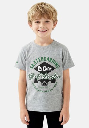 Garçon blond souriant, portant un t-shirt gris avec un motif de skateboard vert et noir et un pantalon noir, debout devant un fond blanc.