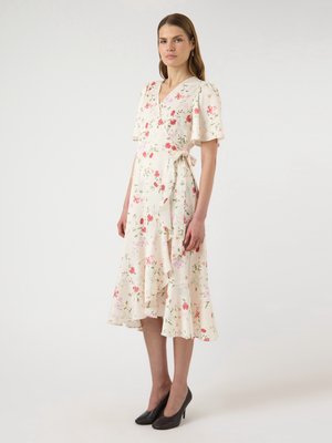 Kvinde iført cremegul slå-om-kjole med pink blomsterprint og flagrende ærmer, kombineret med sorte sko med hæl, stående mod en ensfarvet baggrund.