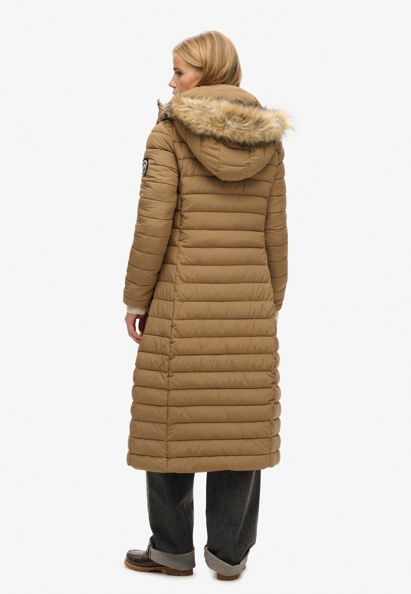 Langer brauner Daunenmantel mit einer mit Fell gefütterten Kapuze, horizontalem Steppmuster und Seitentaschen, getragen mit grauen Hosen und braunem Schuhwerk.