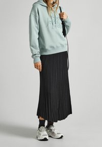 Felpa verde menta con cappuccio e testo in rilievo, abbinata a una gonna lunga plissettata nera e sneakers grigie. Dettagli di calzini neri visibili.