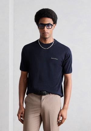 Άνδρας που φοράει σκούρο μπλε μπλουζάκι Marc O'Polo, μπεζ παντελόνι, μαύρη ζώνη, ασημένιο μενταγιόν και μεγάλα σκουρόχρωμα γυαλιά ηλίου, στέκεται μπροστά σε ένα μονόχρωμο φόντο.