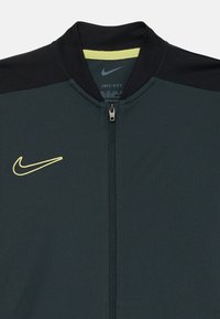 Nike Dri-Fit-jacka i mörk teal med svarta detaljer, ståkrage, hel dragkedja och en framträdande gul logotyp på bröstet. Strukturerad tyg.