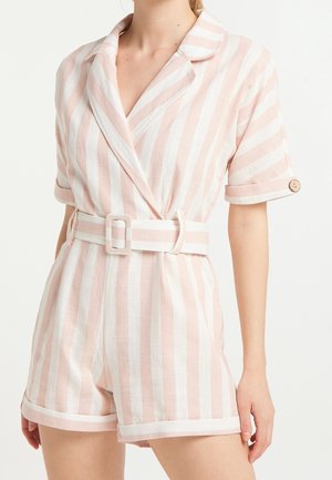 Femme portant une combinaison-short à manches courtes rayée verticalement rose et blanche avec un col, une ceinture en tissu et des poignets retroussés avec un détail de bouton.