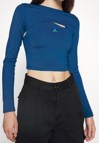Modrý dlouhý rukáv crop top z pružného materiálu, s výřezem a zeleným logem ve středu, kombinovaný s černými kalhotami s vysokým pasem.