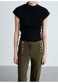 Vrouw in een mouwloze zwarte coltrui en hoogwaist olijfgroene broek met twee rijen decoratieve zilveren knopen.