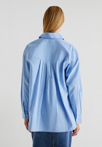 Camicia a righe blu e bianche con dettaglio di piega sulla schiena e maniche lunghe, realizzata in tessuto leggero e morbido.