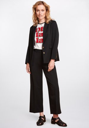 Zwarte op maat gemaakte blazer met gouden knopen, gecombineerd met een witte graphic t-shirt en zwarte wijde broek, aangevuld met zwarte sandalen met bandjes.