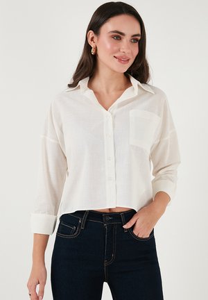 White cropped shirt in lightweight fabric with a button front, long sleeves rolled up, and a single chest pocket. Worn with dark jeans.