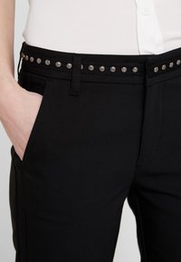 Osoba ubrana w czarne spodnie z posrebrzanym paskiem z ćwiekami, biała koszula włożona w spodnie, ręka włożona w lewą kieszeń.