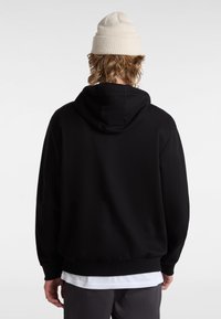 Person mit langen Haaren, trägt eine beige gestrickte Mütze, schwarzen Kapuzenpullover, weißes Shirt darunter und dunkle Hose, steht mit dem Rücken zur Kamera vor weißem Hintergrund.