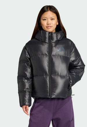 Junge Frau trägt eine schwarze Steppjacke mit hohem Kragen und violette Hose, steht mit einer Hand in der Tasche vor einfarbigem Hintergrund.