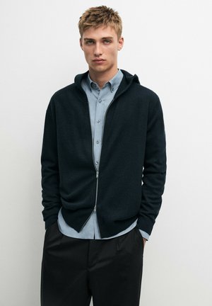 Giovane uomo con le mani in tasca, indossa una camicia blu con bottoni sotto una felpa nera con zip e pantaloni neri su uno sfondo bianco semplice.
