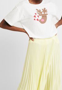 Camiseta blanca con un gráfico de reno marrón con cuernos y nariz rosa, complementada con acentos de corazones rojos. Combinada con una falda amarilla con pliegues.