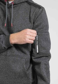 Hoodie gris texturé avec des cordons de serrage accentués en rouge. Il dispose d'une poche à glissière sur la manche et d'une grande poche frontale.