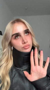 Svart läderjacka med hög krage, långt vågigt blont hår och hand höjd i en gest. Märkbara långa, välmanikyrerade naglar med mörka spetsar.