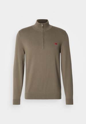 Olivazöld, cipzáras garbó pulóver bordás kötött anyagból. Hosszú ujjakkal, magas gallérral és egy kicsi piros márkalogóval rendelkezik a mellen.