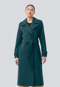 Woman wearing a long dark green double-breasted wool coat with a belt, standing against a plain gray background.