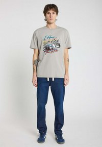 T-shirt graphique en coton gris clair avec impression de course colorée et texte. Associé à un pantalon en denim bleu foncé et des sneakers bleues.