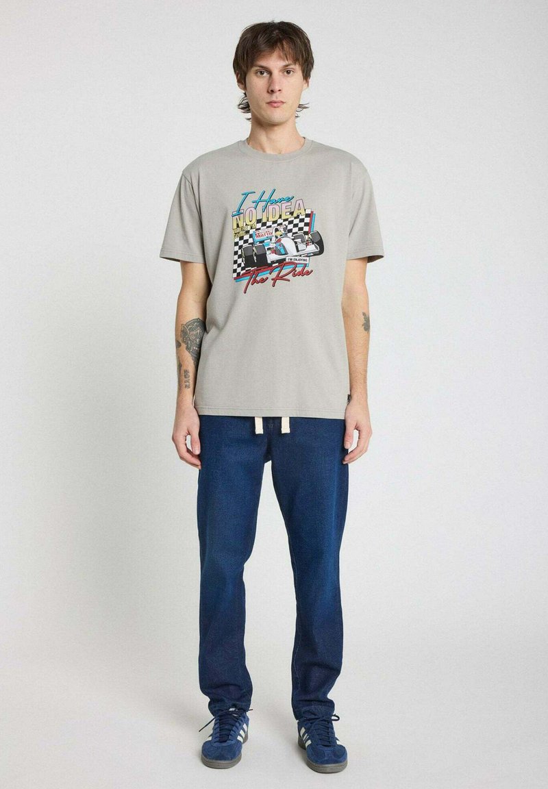 T-shirt graphique en coton gris clair avec impression de course colorée et texte. Associé à un pantalon en denim bleu foncé et des sneakers bleues.