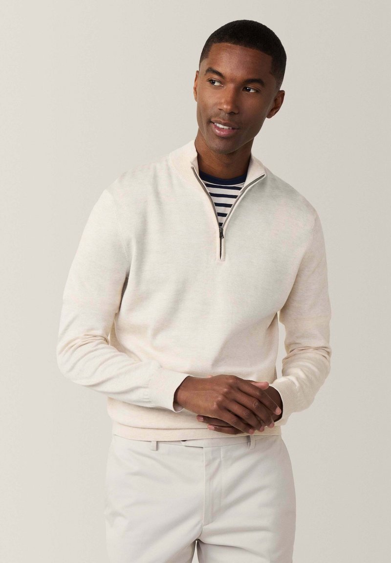 Helle beige Zip-Hoodie mit langen Ärmeln, weichem Stoff und gerippten Bündchen. Trägt man über einem navy- und weißen gestreiften Hemd, kombiniert mit hellen Hosen.