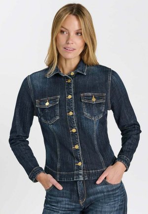 Donna che indossa una giacca di jeans aderente blu scuro con bottoni dorati e due tasche sul petto, con le mani nelle tasche dei jeans abbinati.