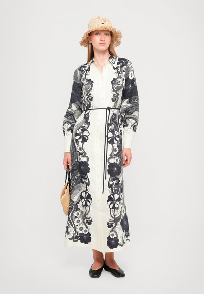 Vestido largo abotonado en crema con patrones oscuros de flores y paisley. Presenta mangas largas, cintura ajustada y falda recta. Se lleva con un sombrero de paja.