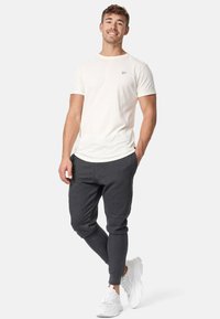 Herrenoutfit, bestehend aus einem cremefarbenen Hemd mit kleinem Logo, dunkelgrauen Jogginghosen und weißen Sneakern. Die Materialien erscheinen weich und bequem.