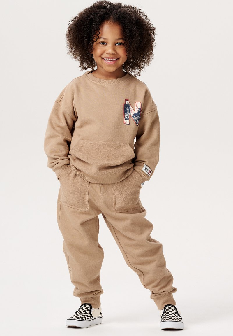 Beige Sweatshirt und Hose mit einer Fronttasche und gesticktem "N"-Logo; weiche Textur; kombiniert mit schwarz-weißen karierten Slippern.