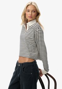 Blonde woman in striped long-sleeve shirt and dark corduroy pants leans on back of wooden chair, looking to her left.