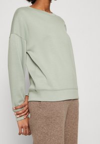 Sudadera verde menta con cuello redondo, mangas largas y un ajuste relajado, hecha de tela suave. Combinada con pantalones marrones texturizados.