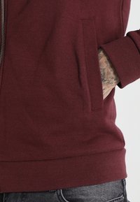 Sudadera burdeos con cremallera, que cuenta con un bolsillo frontal y puños acanalados. Textura suave y diseño de color sólido, combinada con jeans oscuros.