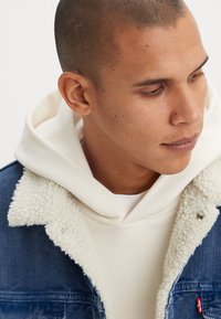 Denim jacket with cream shearling collar, worn over a white hoodie. Features blue ribbed texture and red logo tag on the left pocket.